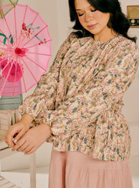 A woman wearing a long-sleeve floral blouse with a gathered peplum waist and a matching dusty pink skirt, standing next to a pink decorative umbrella.