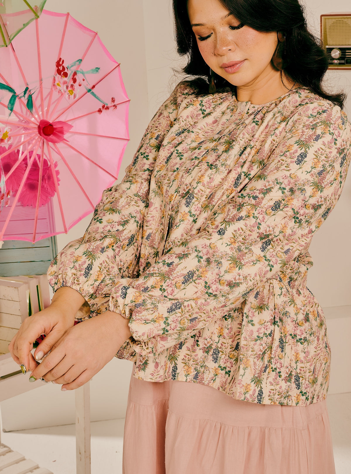 A woman wearing a long-sleeve floral blouse with a gathered peplum waist and a matching dusty pink skirt, standing next to a pink decorative umbrella.