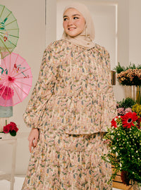 A woman wearing a beige hijab and a long-sleeve, tiered floral modest dress with a matching skirt, standing in a decorated indoor setting with flowers and paper umbrellas.