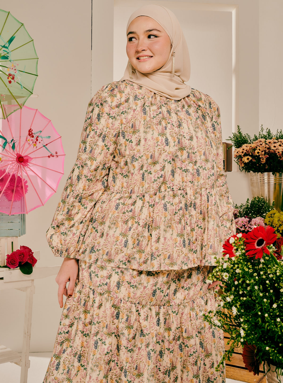 A woman wearing a beige hijab and a long-sleeve, tiered floral modest dress with a matching skirt, standing in a decorated indoor setting with flowers and paper umbrellas.