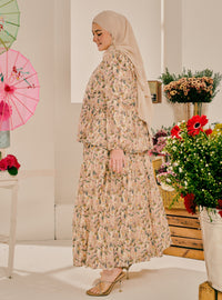 A woman wearing a beige hijab and a long-sleeved, tiered maxi dress with a delicate floral print in soft pink, beige, and green tones. She is standing in a bright, white studio decorated with potted flowers and traditional paper umbrellas.