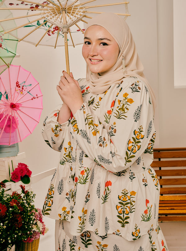 A woman wearing a beige hijab and a cream-colored long-sleeve tunic with a colorful floral and botanical print, holding a decorative paper umbrella. She is posed in an indoor setting with a pink umbrella and flowers in the background.
