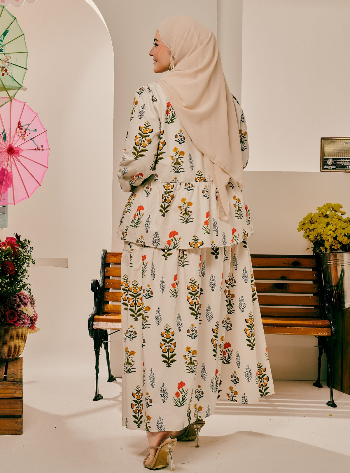 A woman wearing a beige hijab and a cream-colored two-piece outfit with a colorful floral print, standing in a decorated indoor setting with wooden benches and hanging umbrellas.