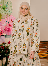 A woman wearing a beige hijab and a long-sleeve tiered abaya dress with a colorful floral print, standing in a decorated indoor setting with pink umbrellas and flowers.