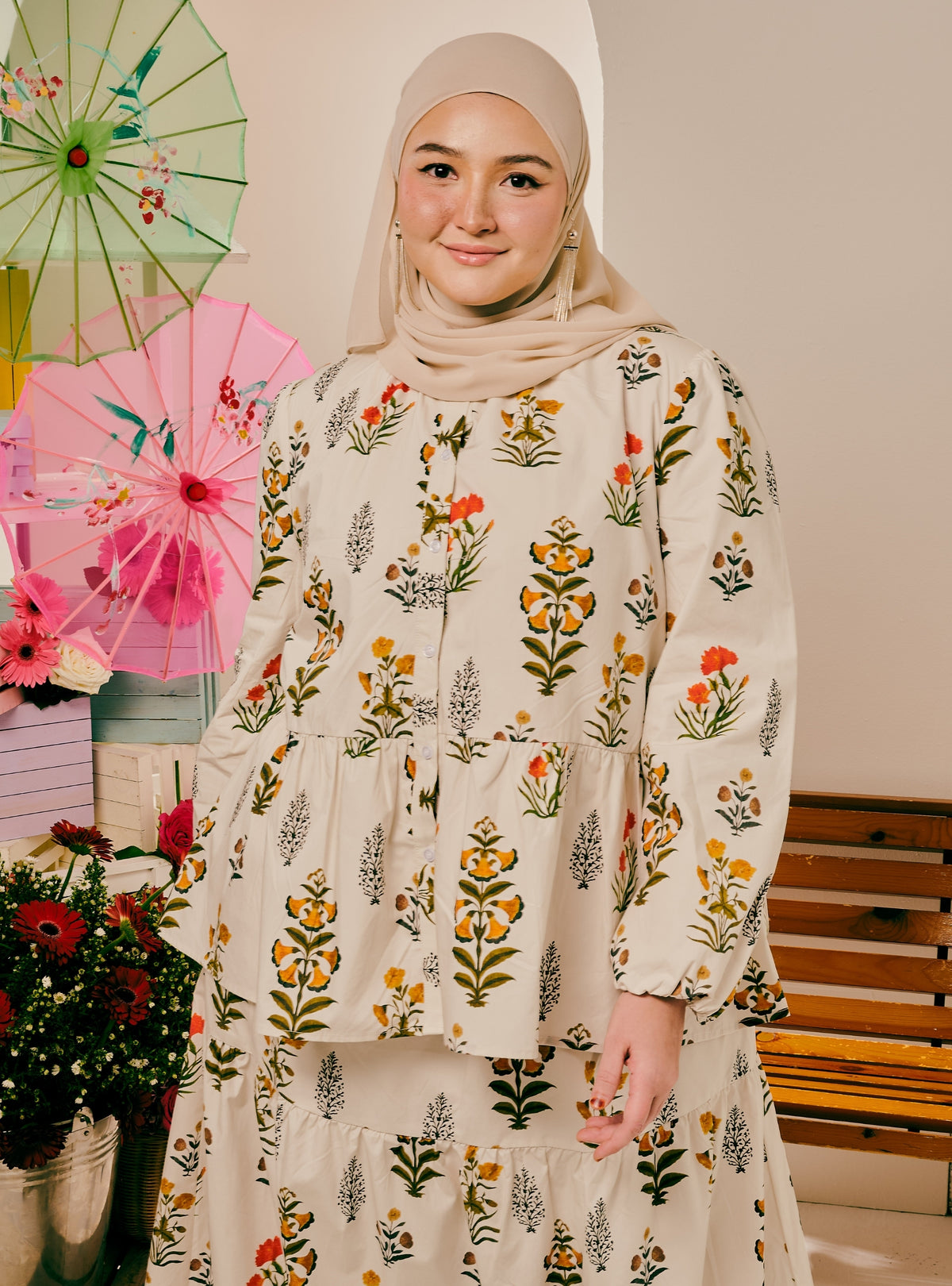 A woman wearing a beige hijab and a long-sleeve tiered abaya dress with a colorful floral print, standing in a decorated indoor setting with pink umbrellas and flowers.