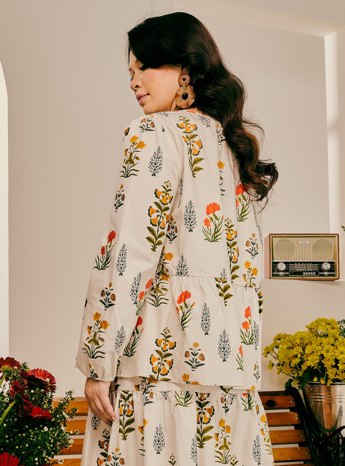 A woman wearing a cream-colored long-sleeve tunic top and matching skirt set, both decorated with a colorful floral and botanical print. She is photographed from behind in a warm, indoor setting with a vintage radio on the wall and flowers in the foreground.