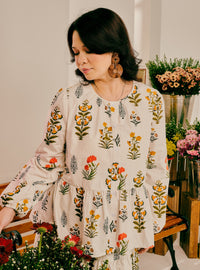 A woman wearing a cream-colored long-sleeve blouse with a colorful floral block print, standing in a flower-filled room.