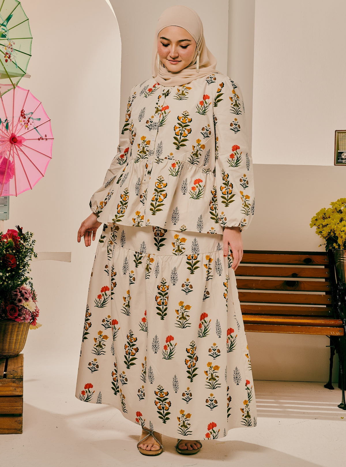 A woman wearing a beige two-piece modest outfit with a floral print, consisting of a long-sleeve peplum top and a matching maxi skirt, paired with a beige hijab. She is standing in a brightly lit indoor setting with decorative umbrellas and a wooden bench in the background.