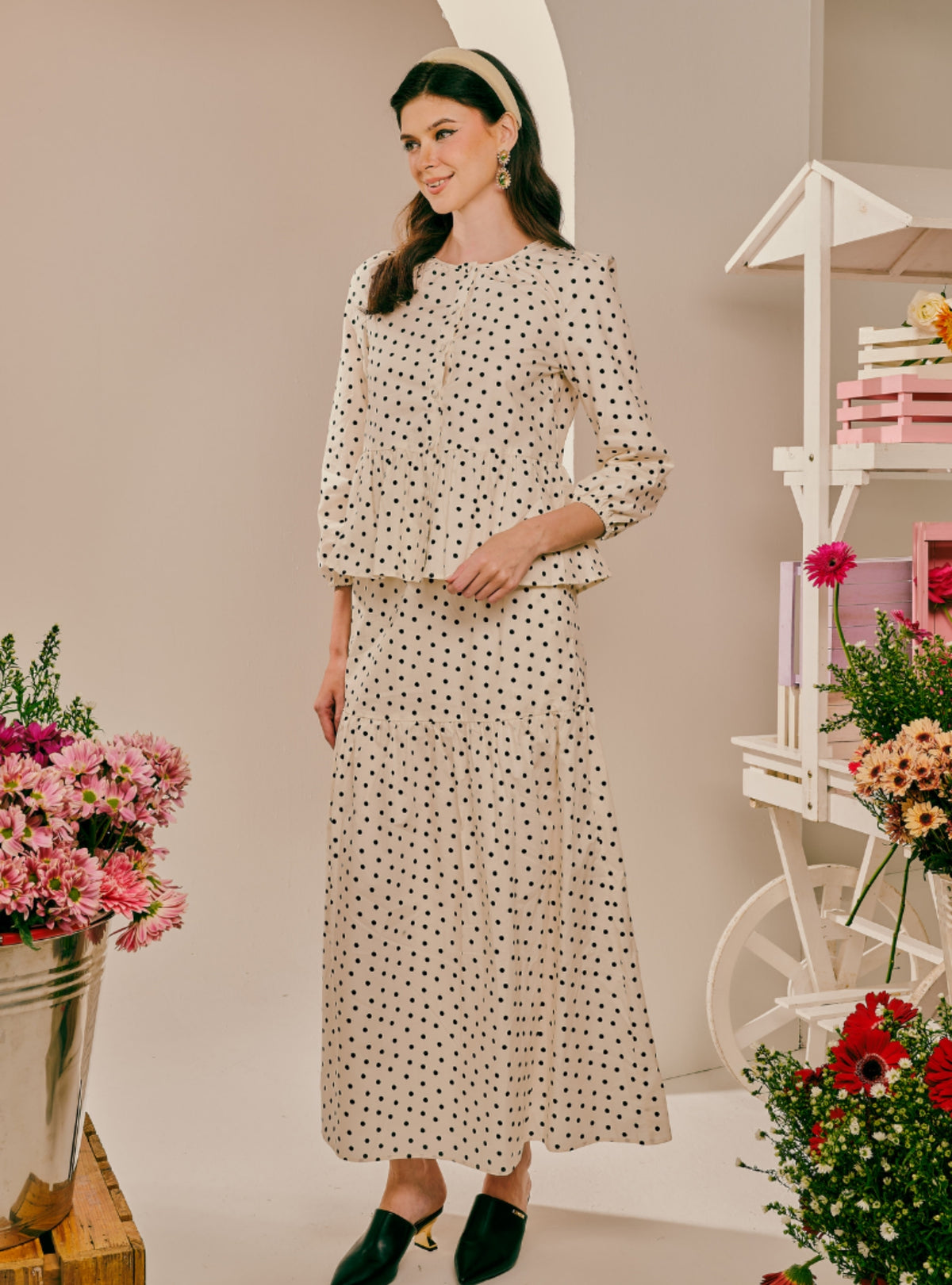 A woman models a matching two-piece long-sleeve dress set with a beige base and black polka dots, styled with black pointed mule heels in a bright, styled studio setting with floral arrangements.