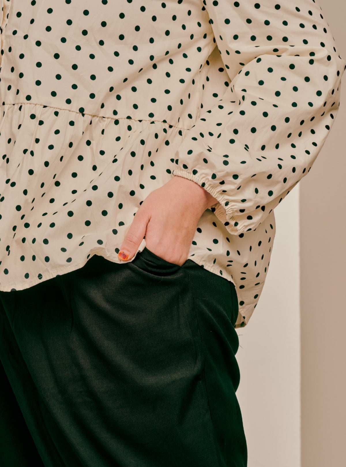 A close-up of a person wearing a cream-colored long-sleeve blouse with a black polka dot pattern and dark green satin-like trousers, with one hand in the pocket of the trousers.