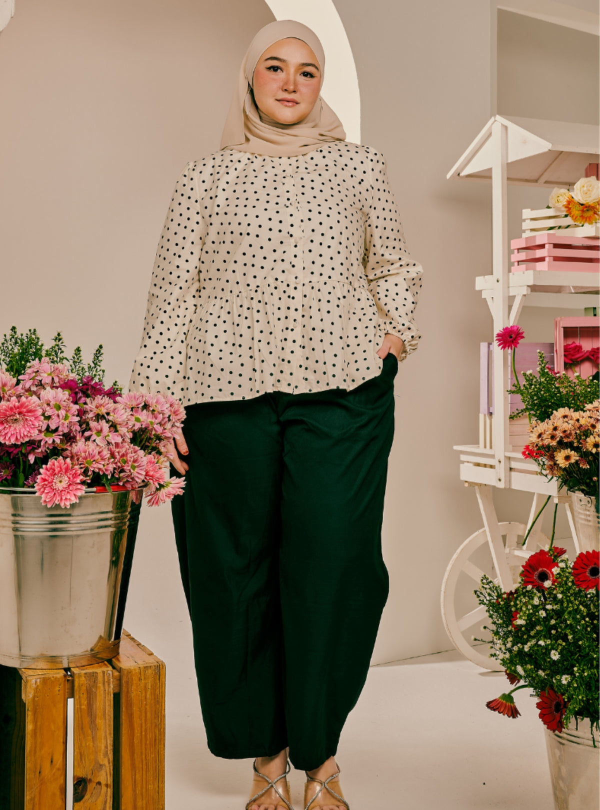 A woman wearing a beige hijab, a cream long-sleeve blouse with black polka dots, and dark green wide-leg pants, standing in a styled indoor setting with flowers and a white decorative cart.
