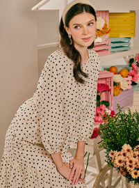 A woman wearing a cream-colored long-sleeve dress with black polka dots, styled with a matching beige beret and dangling earrings, posed in a brightly decorated room with flowers and pastel shelves.