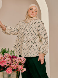 A woman wearing a beige hijab and a cream long-sleeve blouse with black polka dots, paired with dark green pants, posing next to a bucket of pink flowers against a beige arched backdrop.