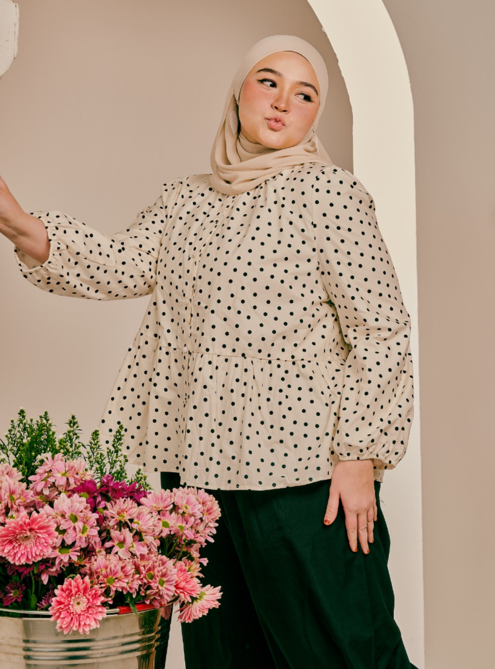A woman wearing a beige hijab and a cream long-sleeve blouse with black polka dots, paired with dark green pants, posing next to a bucket of pink flowers against a beige arched backdrop.