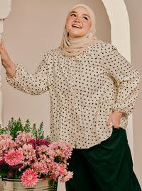 A woman wearing a beige hijab and a cream-colored long-sleeve blouse with black polka dots, paired with dark green trousers. She is standing in front of a beige arched wall with a bucket of pink flowers in the foreground.