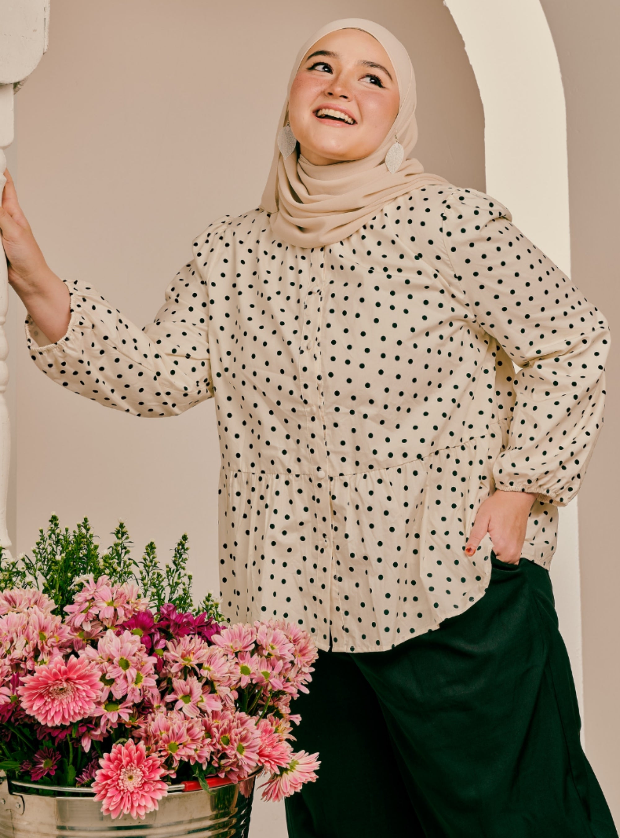 A woman wearing a beige hijab and a cream-colored long-sleeve blouse with black polka dots, paired with dark green trousers. She is standing in front of a beige arched wall with a bucket of pink flowers in the foreground.