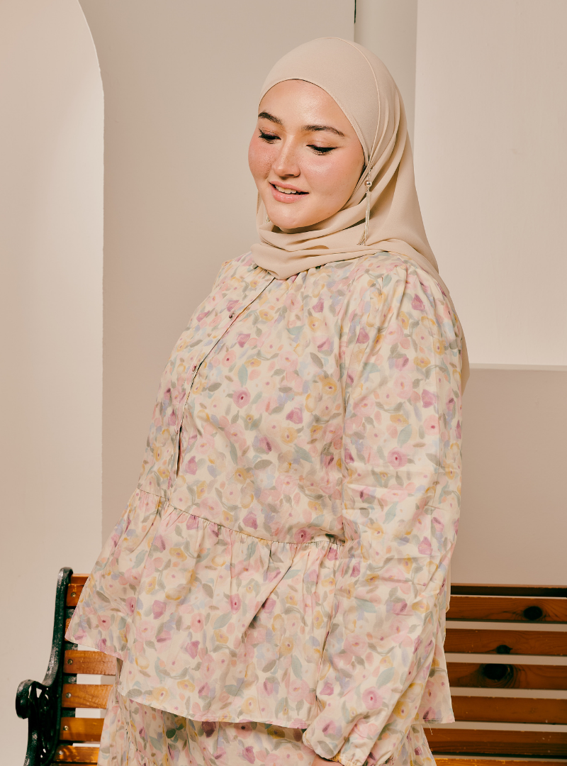 A woman wearing a beige hijab and a long-sleeve floral print abaya with a peplum waist, standing in an indoor setting.