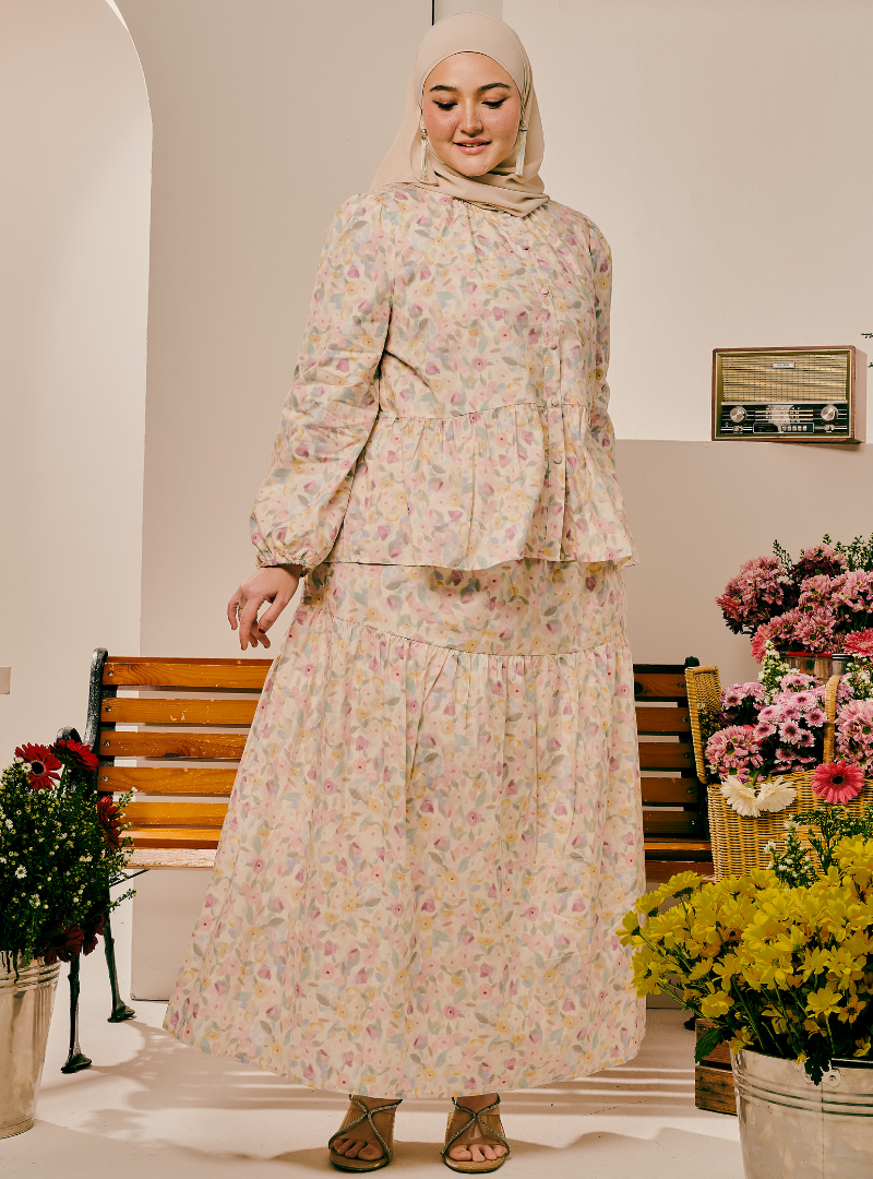 A woman wearing a two-piece modest outfit with a long-sleeve peplum top and a matching tiered maxi skirt, both in a light beige floral print. She is also wearing a light beige hijab and brown lace-up sandals, standing in an indoor setting decorated with flowers and wooden benches.