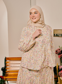 A woman wearing a beige hijab and a matching long-sleeve tiered tunic dress with a delicate pastel floral print, standing in an indoor setting with a wooden bench and flowers in the background.