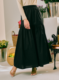 A black tiered maxi skirt worn by a model, styled with a beige long-sleeve top and clear high-heeled sandals, set against a bright indoor background with decorative baskets and plants.