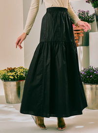 A woman wearing a black tiered maxi skirt with a smocked waistband, paired with a beige long-sleeve top and clear high-heeled sandals, standing in a bright indoor setting with potted plants.