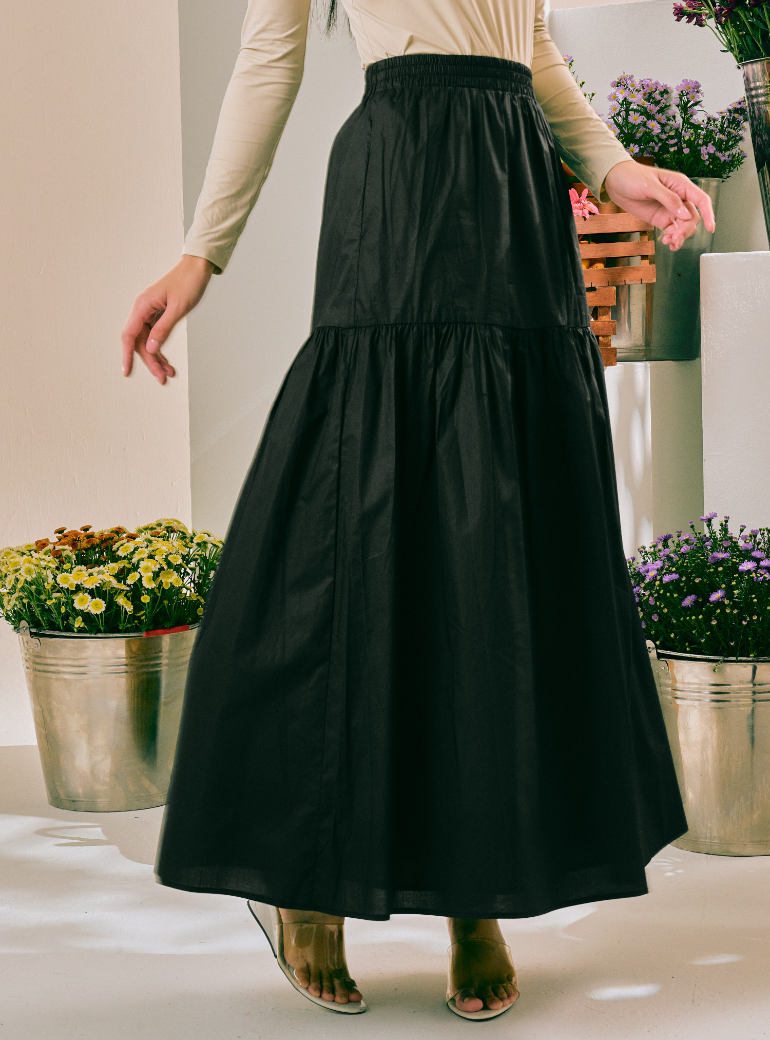 A woman wearing a black tiered maxi skirt with a smocked waistband, paired with a beige long-sleeve top and clear high-heeled sandals, standing in a bright indoor setting with potted plants.