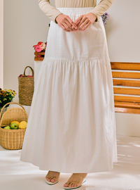 A woman wearing a long, tiered white maxi skirt with a smocked waistband, paired with a beige long-sleeve top and flat sandals, standing in a bright indoor setting with wicker baskets and a wooden bench in the background.