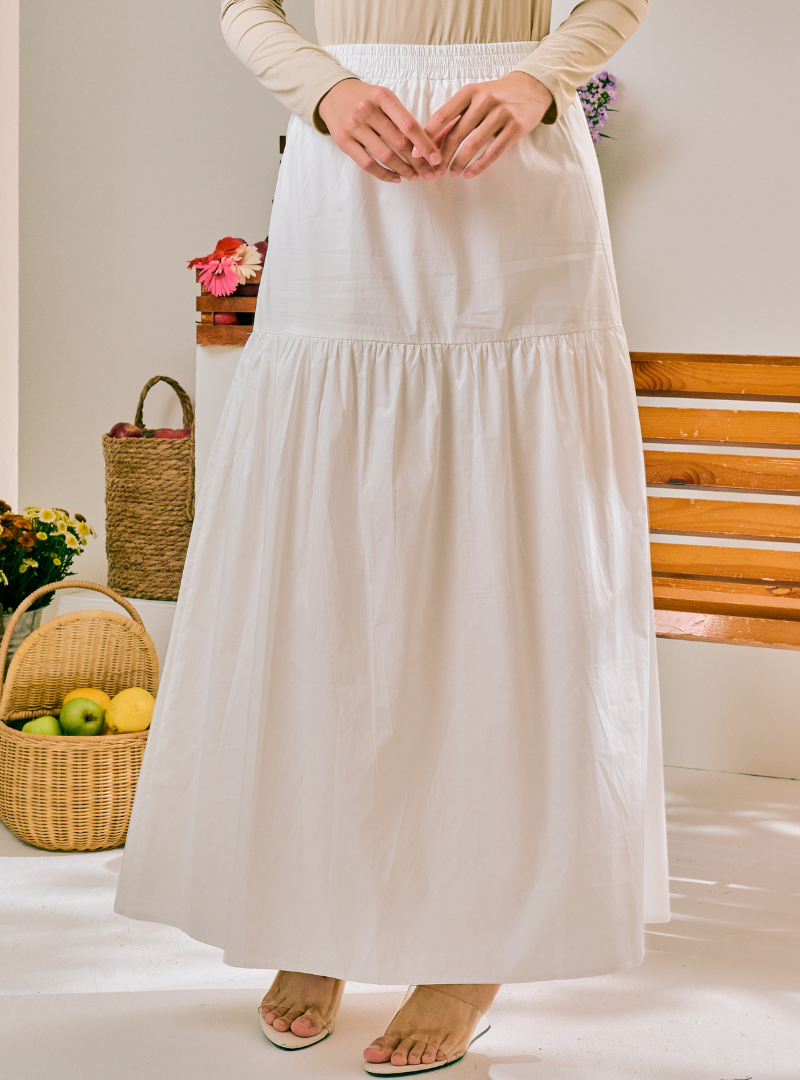 A woman wearing a long, tiered white maxi skirt with a smocked waistband, paired with a beige long-sleeve top and flat sandals, standing in a bright indoor setting with wicker baskets and a wooden bench in the background.