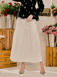A model wearing a black long-sleeve top with white floral embroidery and a high-waisted, tiered white midi skirt, styled with silver heeled sandals. The background features rustic wooden crates holding potted red and pink flowers.
