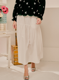 A model wearing a cream-colored tiered midi skirt paired with a black velvet long-sleeve top with white butterfly prints and silver sandals, standing in a brightly lit indoor setting.
