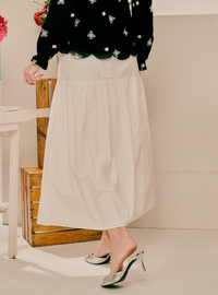 A model wearing a long, tiered off-white midi skirt paired with a black floral long-sleeve top and silver mule heels, standing in a studio setting.