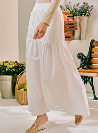 A woman wearing a long white tiered maxi skirt with a beige long-sleeve top and clear heeled sandals, standing in a bright indoor setting with potted plants and a bench in the background.