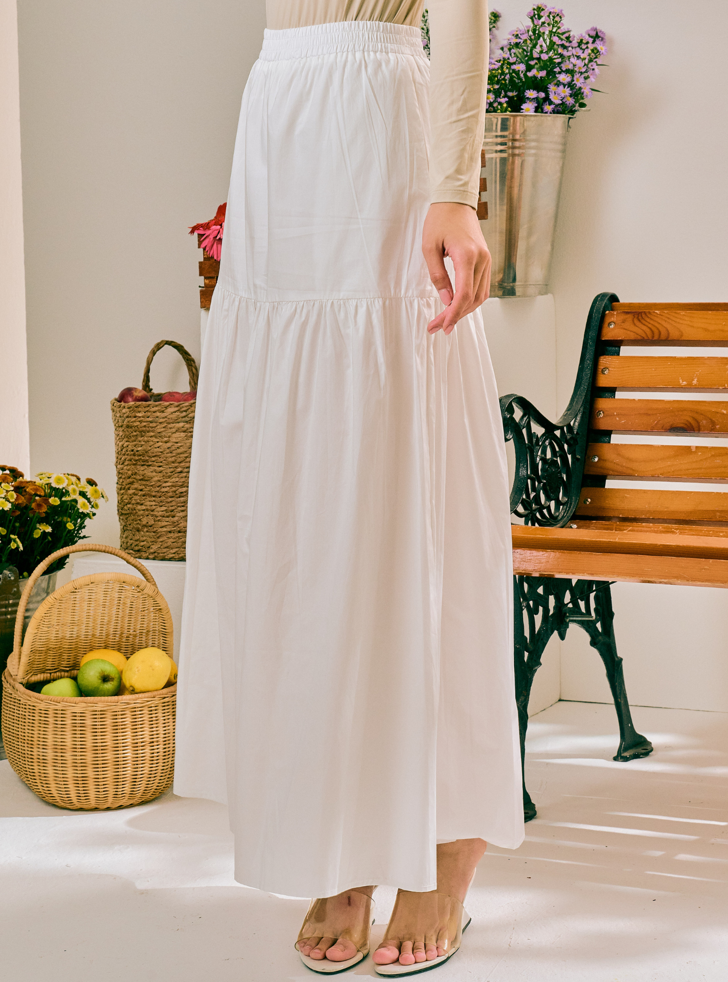 A woman wearing a long white tiered maxi skirt with a beige ribbed top, standing in a bright indoor setting with a wooden bench, wicker baskets, and flowers in the background.
