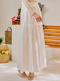 A woman wearing a long white tiered maxi skirt with a cream long-sleeve top, standing in a bright indoor setting with a wooden bench and woven baskets in the background.