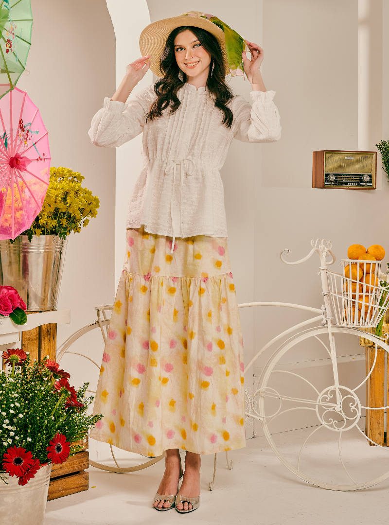 A woman stands in a styled indoor set wearing a cream pleated long-sleeve blouse and a matching tiered maxi skirt with a yellow, pink, and orange floral print. She is holding a wide-brimmed straw hat and posing in front of a white decorative bicycle with flowers and umbrellas.