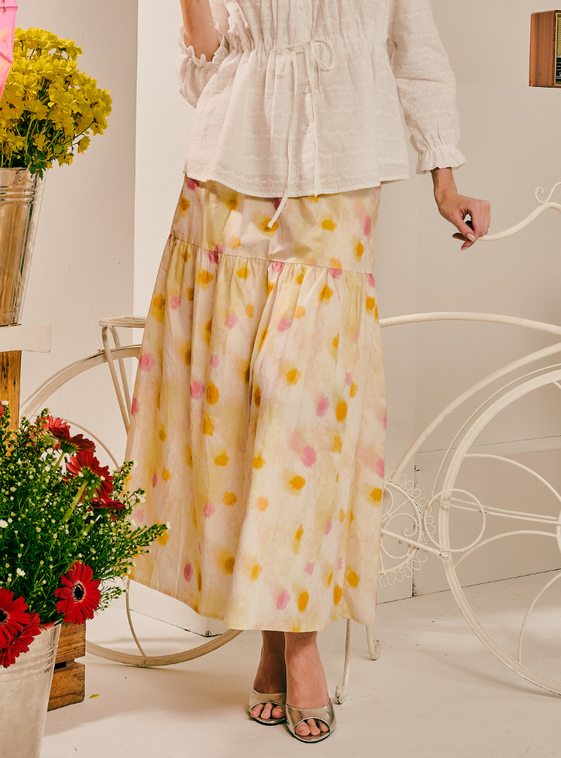 A woman wearing a cream-colored tiered maxi skirt with yellow and pink watercolor-style polka dots, paired with a matching cream top. She is standing in a styled indoor setting with white decorative bicycle frames and potted flowers.