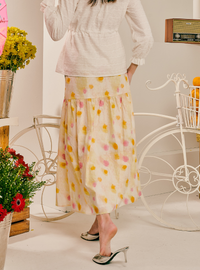 A woman wearing a cream-colored tiered midi skirt with a yellow and pink floral print, paired with a white long-sleeve blouse and silver heels, standing in a styled room with white decorative bicycles and flowers.