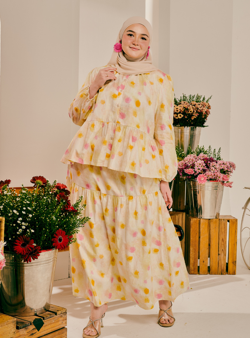 A woman wearing a beige hijab and a matching tiered long-sleeve maxi dress with a pink and yellow floral print, standing in a styled studio setting with potted flowers.