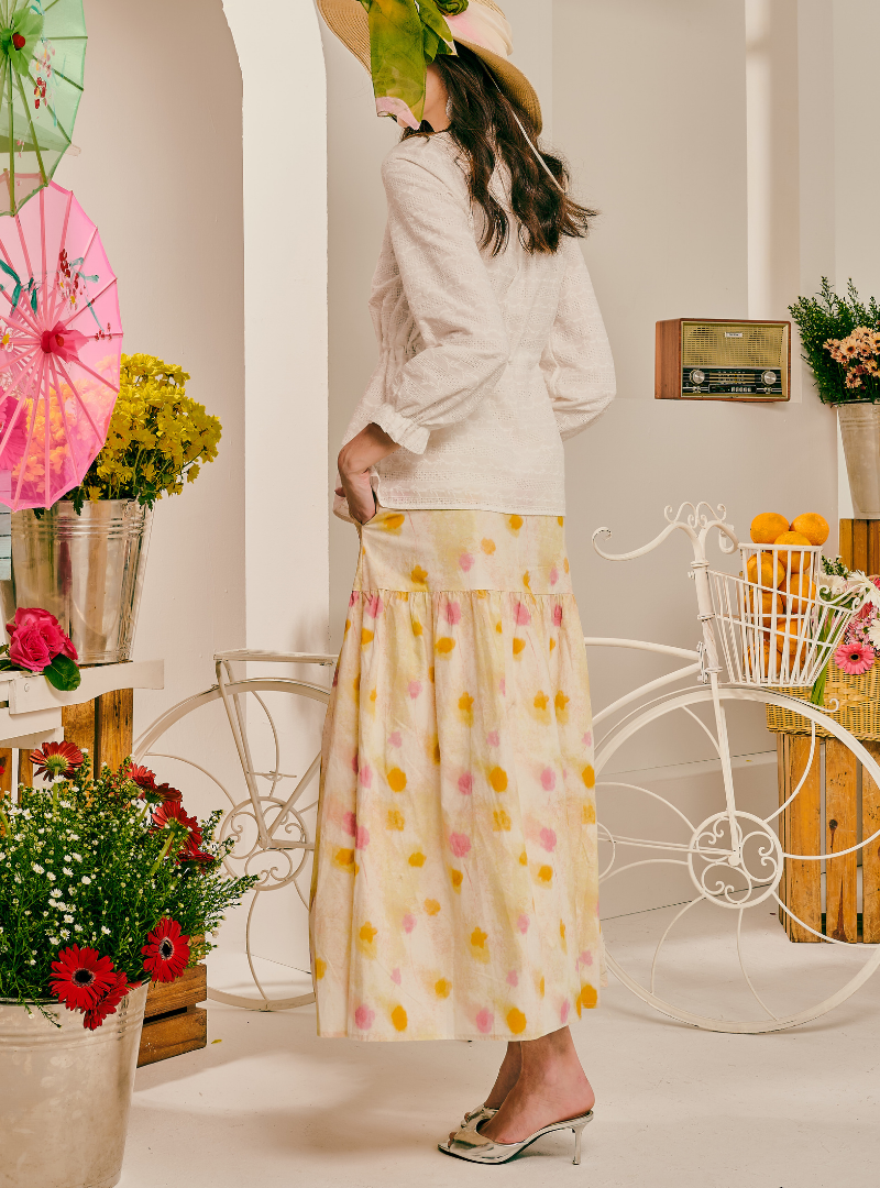 A woman stands in a styled indoor set wearing a cream long-sleeve wrap-style top and a matching cream maxi skirt with pink and yellow floral print. She is seen from the back, wearing a straw hat and metallic heeled sandals.