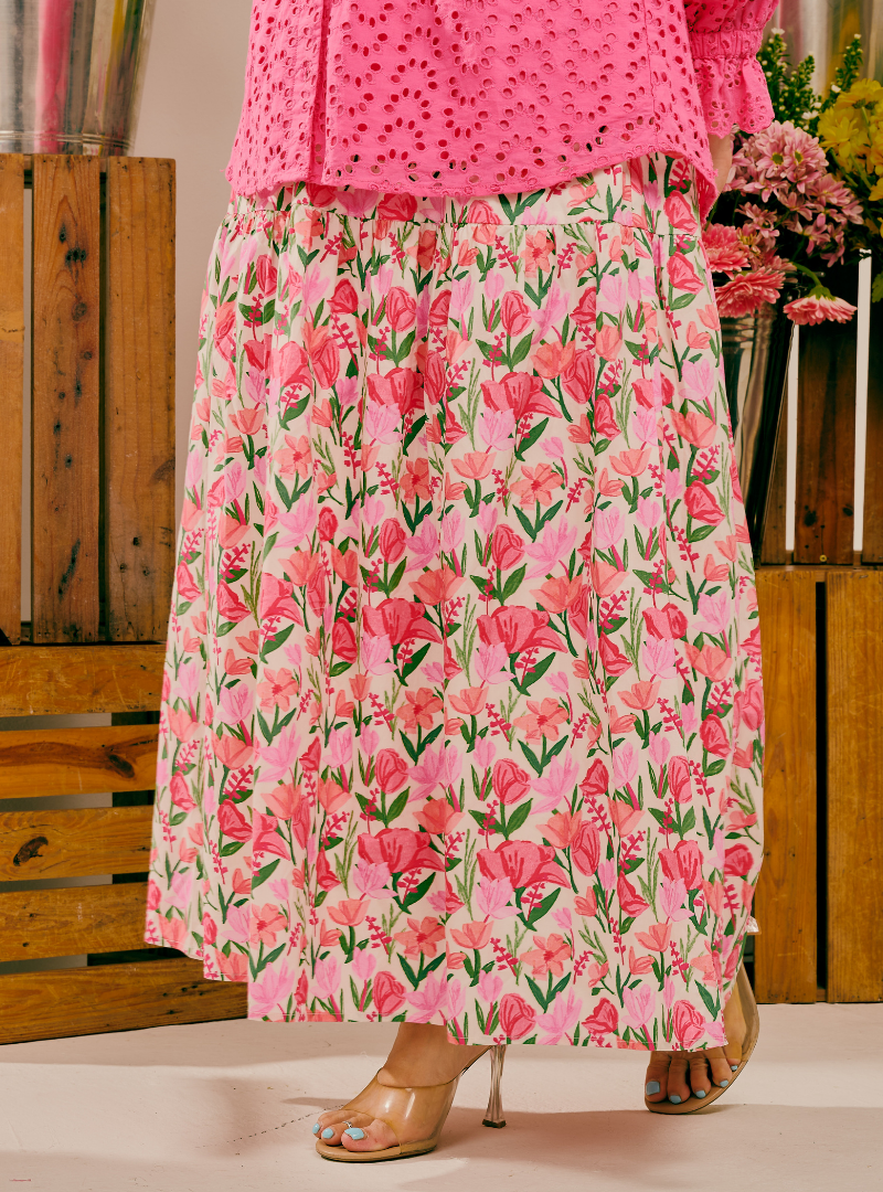A person wearing a bright pink eyelet top and a long pink skirt with a dense floral print in pink, red, and green, paired with clear high-heeled sandals, standing in front of a rustic wooden crate background.