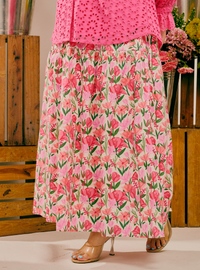 A person wearing a bright pink eyelet top and a long pink skirt with a dense floral print in pink, red, and green, paired with clear high-heeled sandals, standing in front of a rustic wooden crate background.