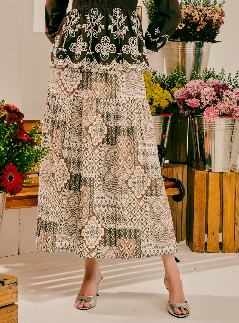 A model wearing a long, tiered patchwork maxi skirt with intricate geometric and floral patterns in beige, brown, and black, paired with a matching embroidered top. The setting is a rustic indoor studio decorated with flower arrangements in metal buckets.