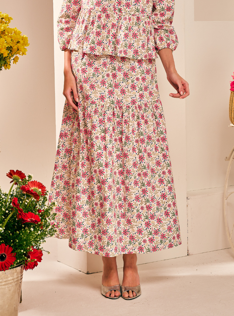 A woman wearing a matching two-piece floral set consisting of a long tiered maxi skirt and a long-sleeve peplum top. The set features a pink and green floral pattern on a cream background, styled with clear high-heeled sandals.