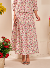 A woman wearing a matching two-piece floral set consisting of a long tiered maxi skirt and a long-sleeve peplum top. The set features a pink and green floral pattern on a cream background, styled with clear high-heeled sandals.