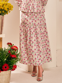 A woman wearing a long cream maxi skirt with a pink and green floral print, paired with a matching ruffled top and metallic silver heeled sandals. The scene is styled with potted red and yellow flowers in the foreground against a cream-colored wall.
