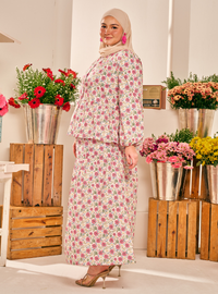 A woman modeling a two-piece floral print set consisting of a long-sleeve top and a matching maxi skirt, paired with a beige hijab and gold heels, set against a background of potted flowers and wooden crates.