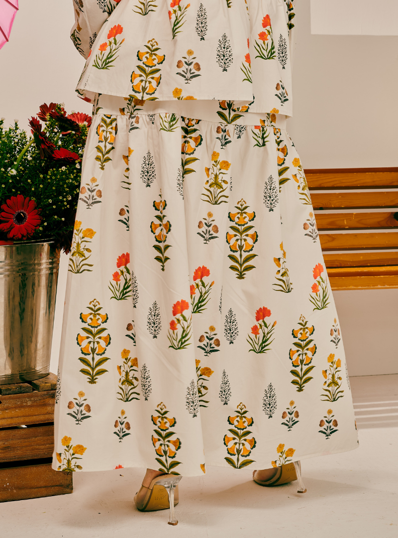 A two-piece matching set featuring a cropped tunic top and a high-waisted maxi skirt, both printed with a colorful floral and botanical pattern on an off-white background. The pattern includes orange, yellow, green, and gray botanical motifs.