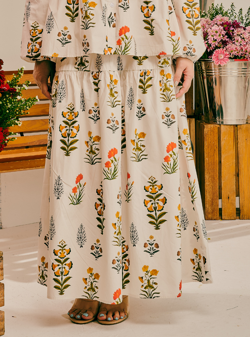 A person wearing a cream-colored two-piece set with a colorful floral print, consisting of a ruffled long-sleeve top and a matching tiered maxi skirt, paired with brown sandals.