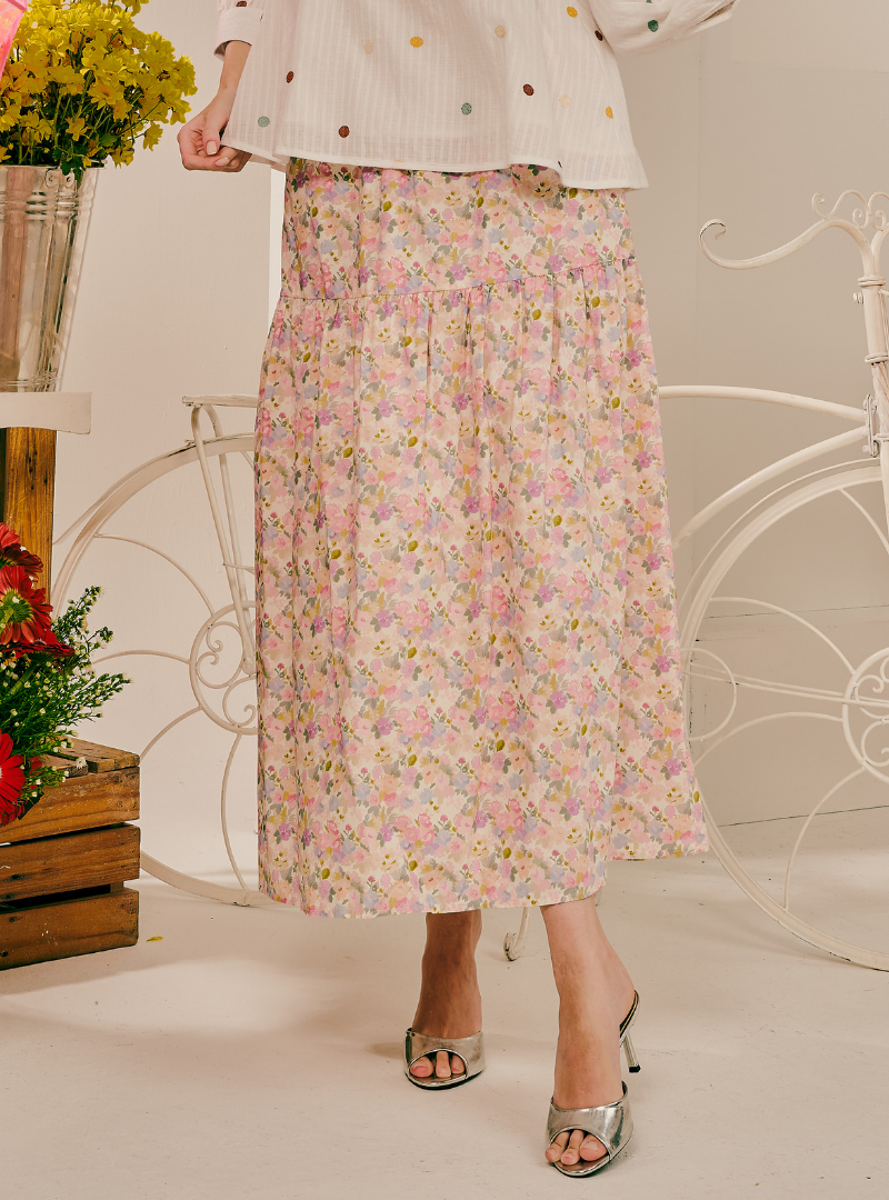 A woman wearing a long, pleated midi skirt with a pastel pink, yellow, and white floral print, styled with a white polka-dot top and metallic silver heeled sandals.