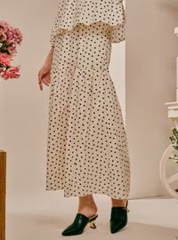 A model wearing a cream-colored maxi dress with a black polka dot pattern and dark green pointed-toe mule heels, shown from the waist down in an indoor setting.